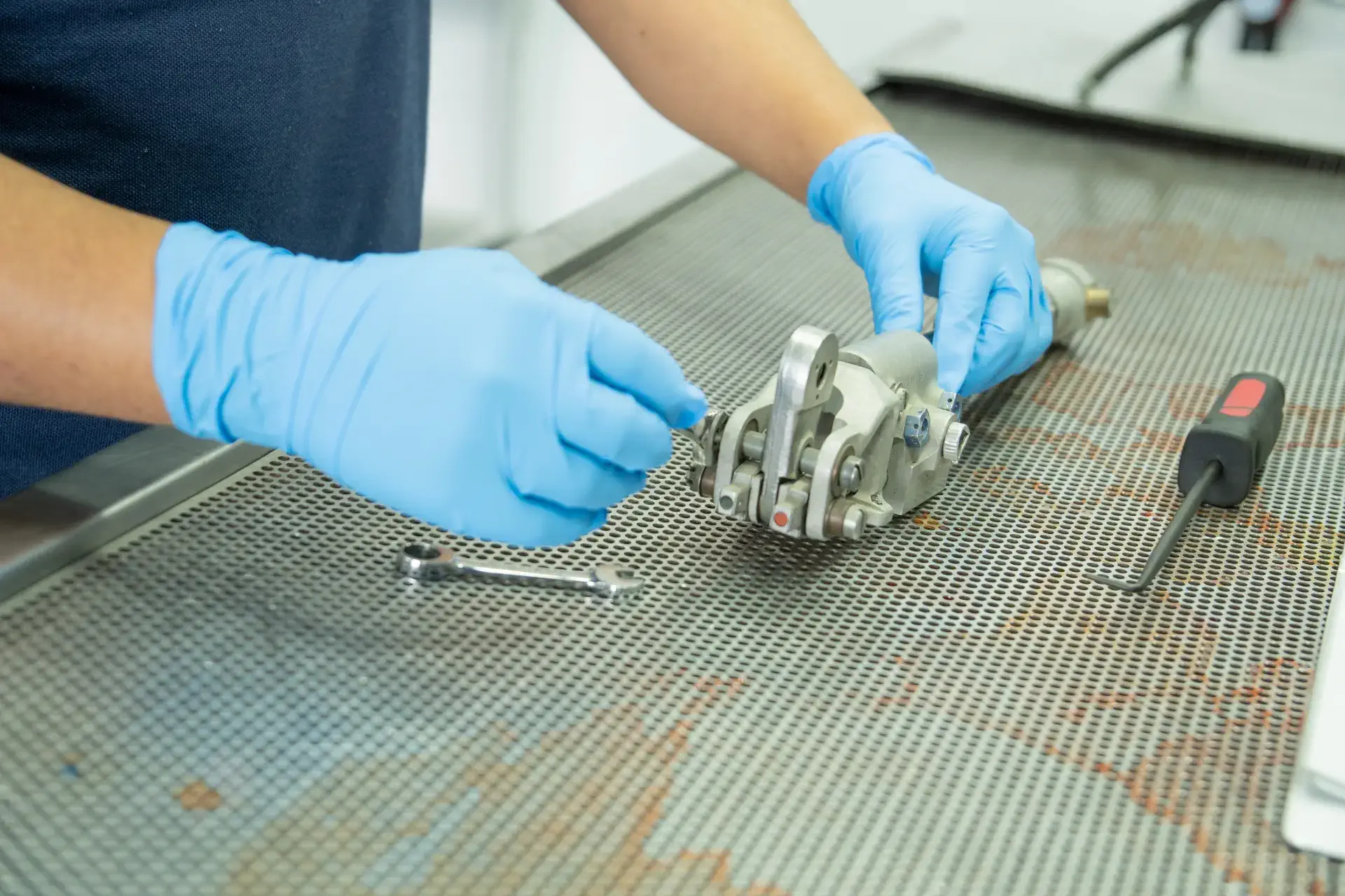 Person in blue gloves repairing a mechanical part on a metal table with tools nearby.