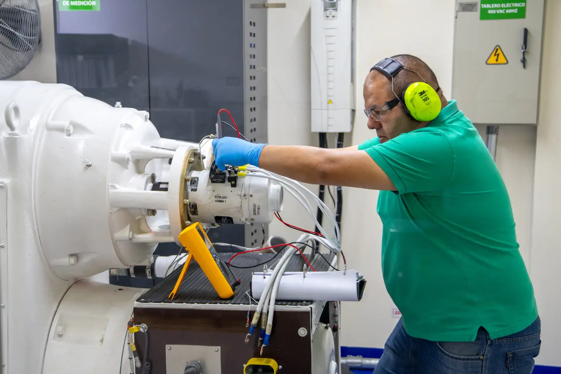 A technician in a green shirt works on industrial machinery with wiring and tools.