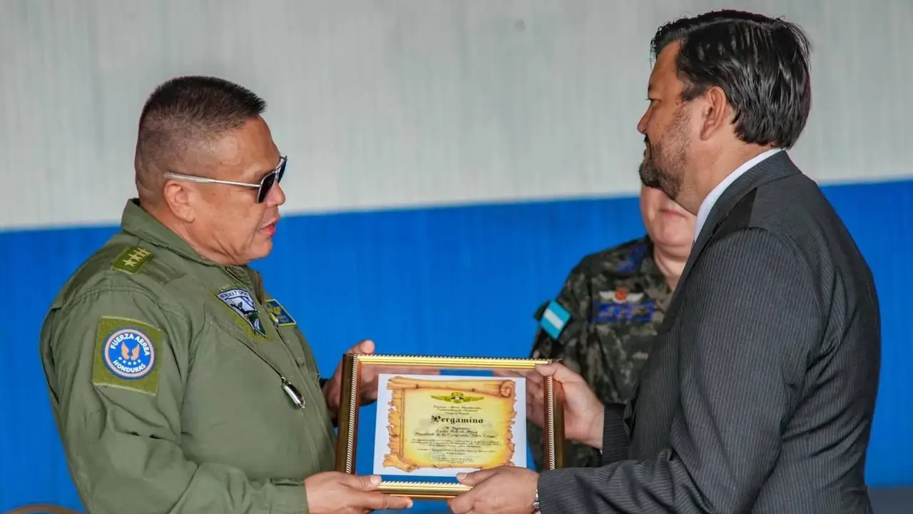 Two individuals exchanging a framed certificate. One is in a military uniform, the other in a suit.